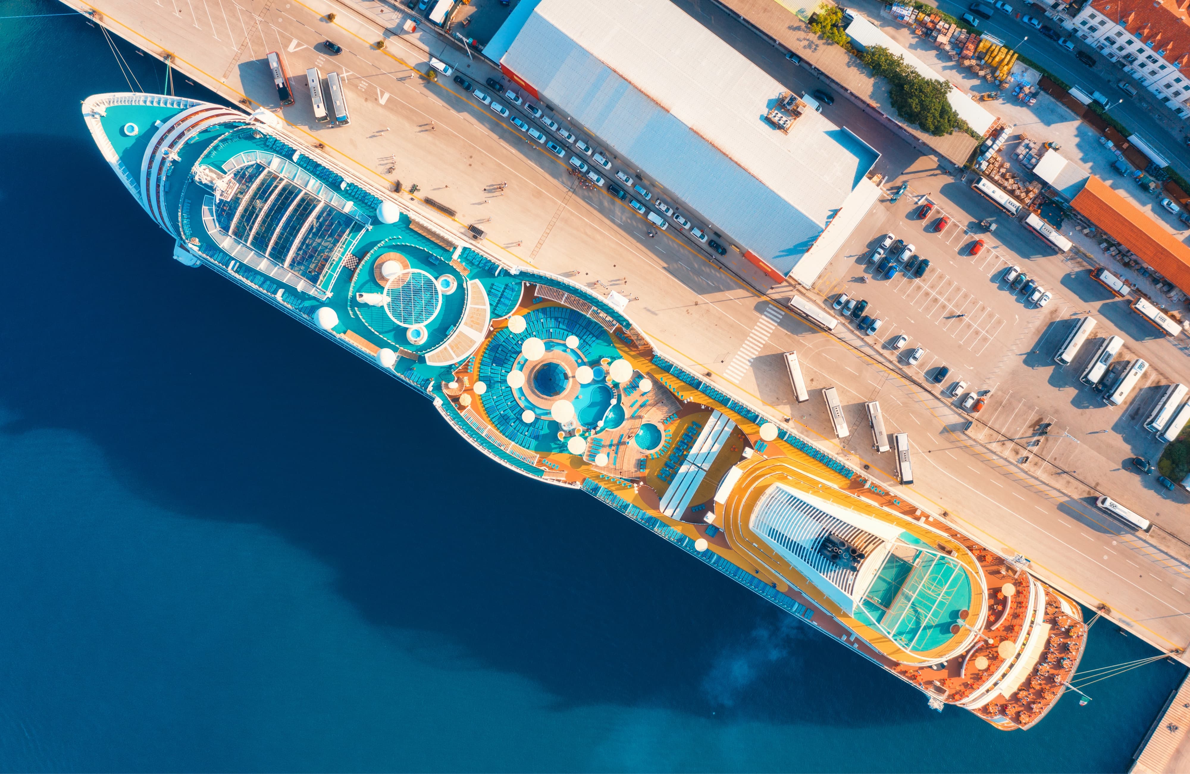 Aerial view of luxury cruise liner in port. Top view of swimming pool with sunbeds, umbrellas and deck chairs, wooden deck on the cruise ship, cars and buildings in summer. View from above