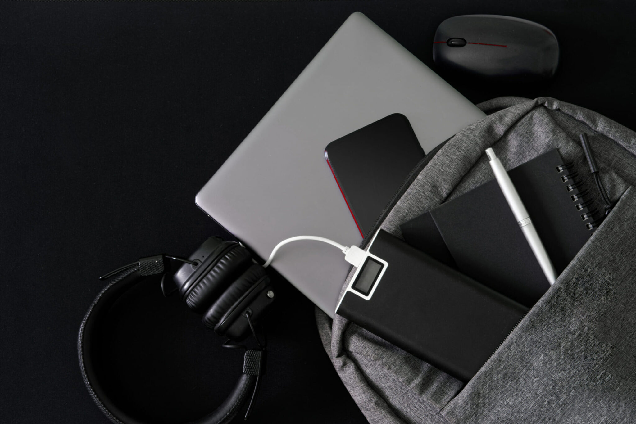Silver laptop and modern gadgets lie in a gray backpack on a dark table