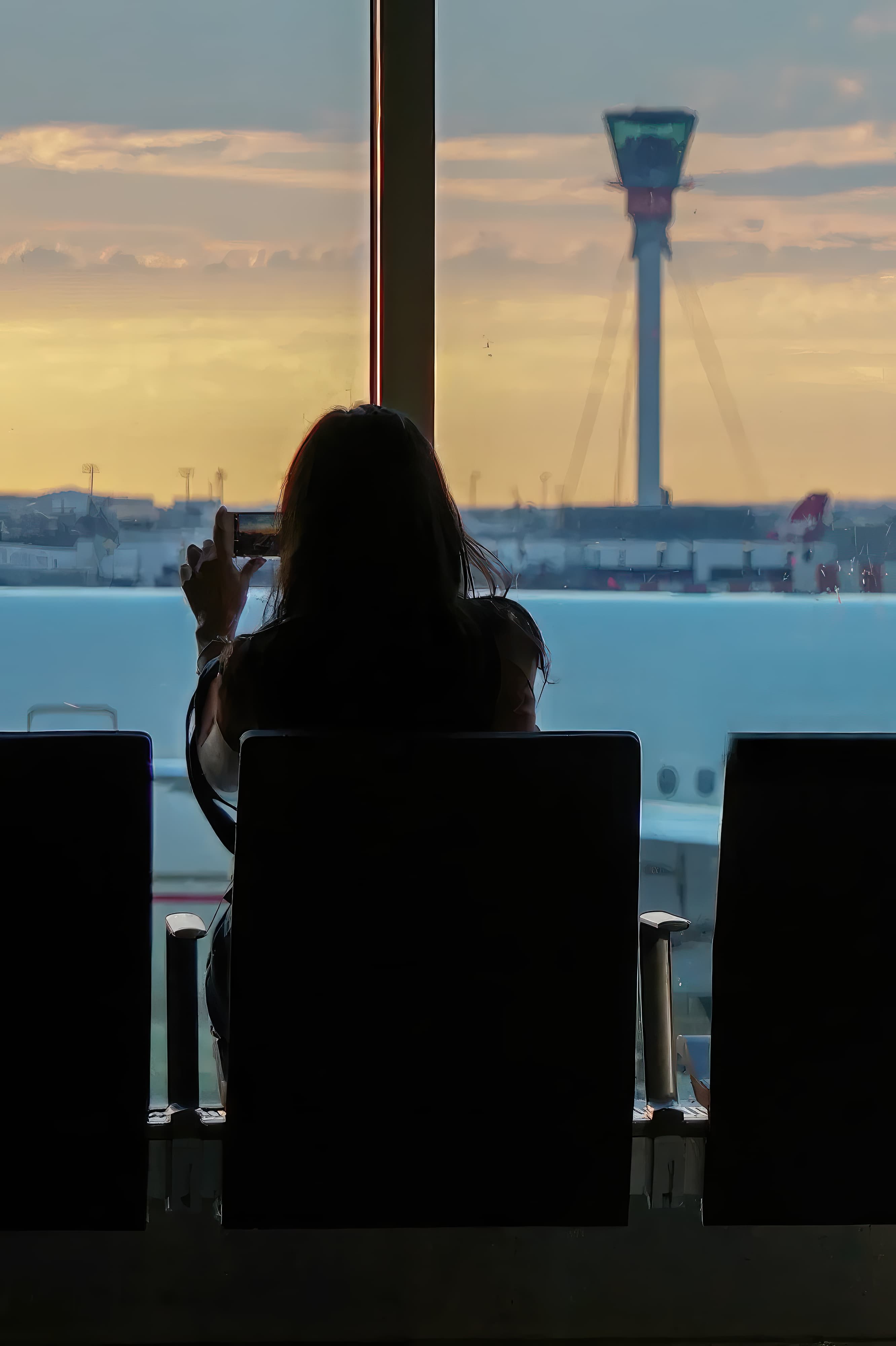 An adult female traveler looking out of an airport window at sunset taking pictures with her smartphone