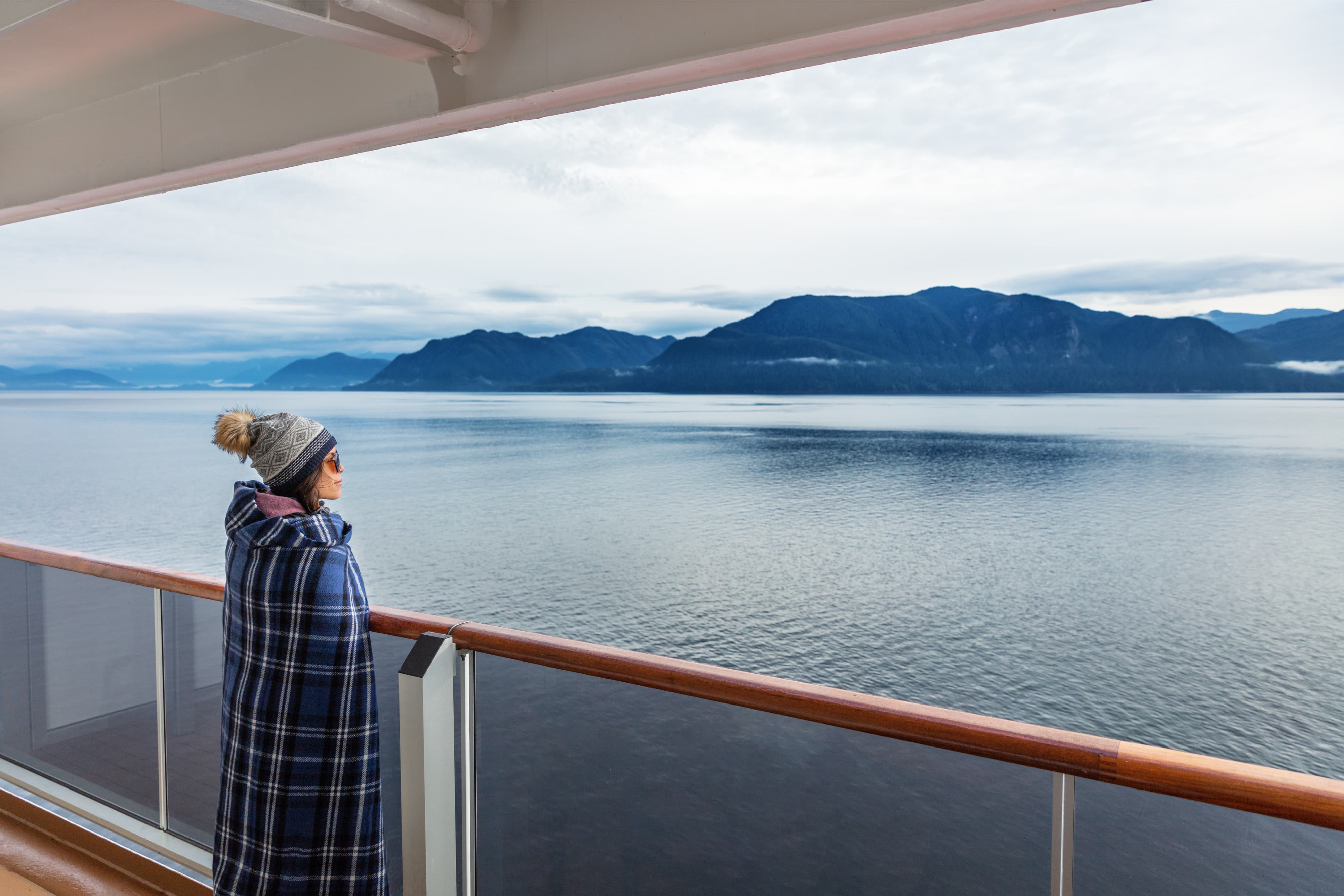 Alaska cruise travel luxury vacation woman watching inside passage scenic cruising day on balcony deck enjoying view of mountains and nature landscape. Asian girl tourist with wool blanket